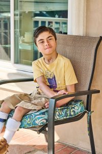 14-year-old boy named Marcos sitting in a cushioned outdoor chair on a brick patio, wearing a yellow graphic t-shirt, khaki shorts, white leg braces, and brown shoes, with a calm expression and short dark hair.